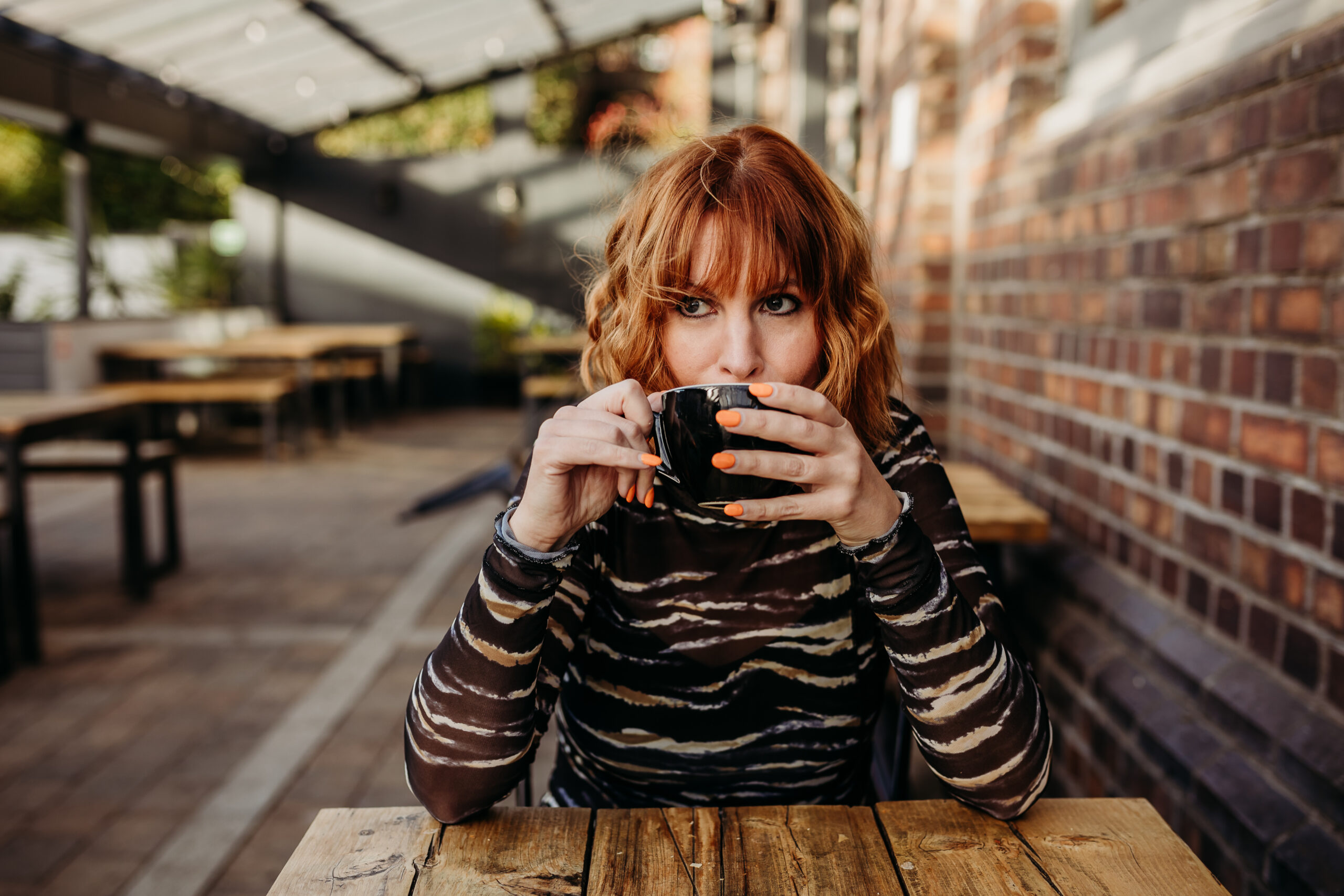 White woman with wavy ginger hair, an animal print dress and orange nails looks off to the side, playfully, while holding a coffee cup in front of her mouth