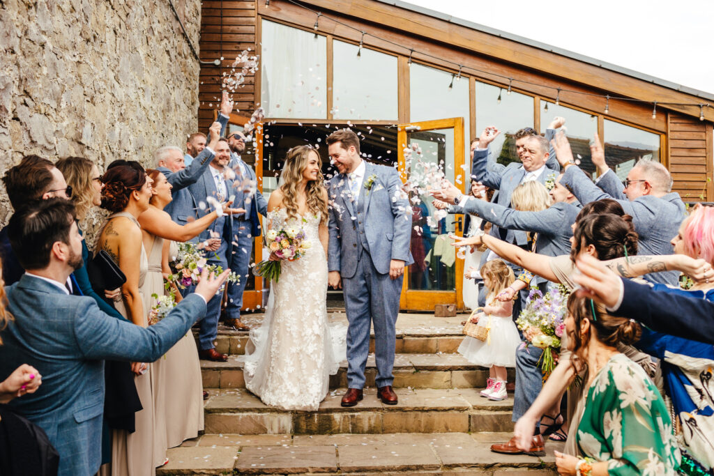 Bride and groom are stood on steps, looking at each other and smiling in anticipation as a confetti shower is about to hit them