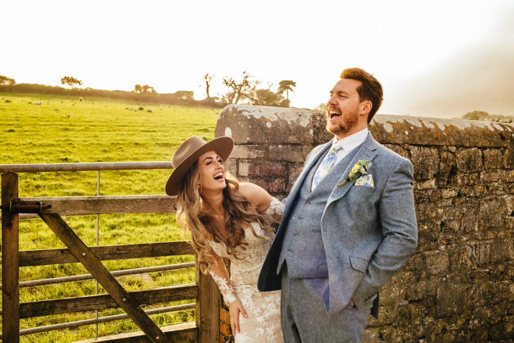 Bride and groom laughing during golden hour. The bride wears a lace dress and cowboy hat, the groom wears a light blue suit. 