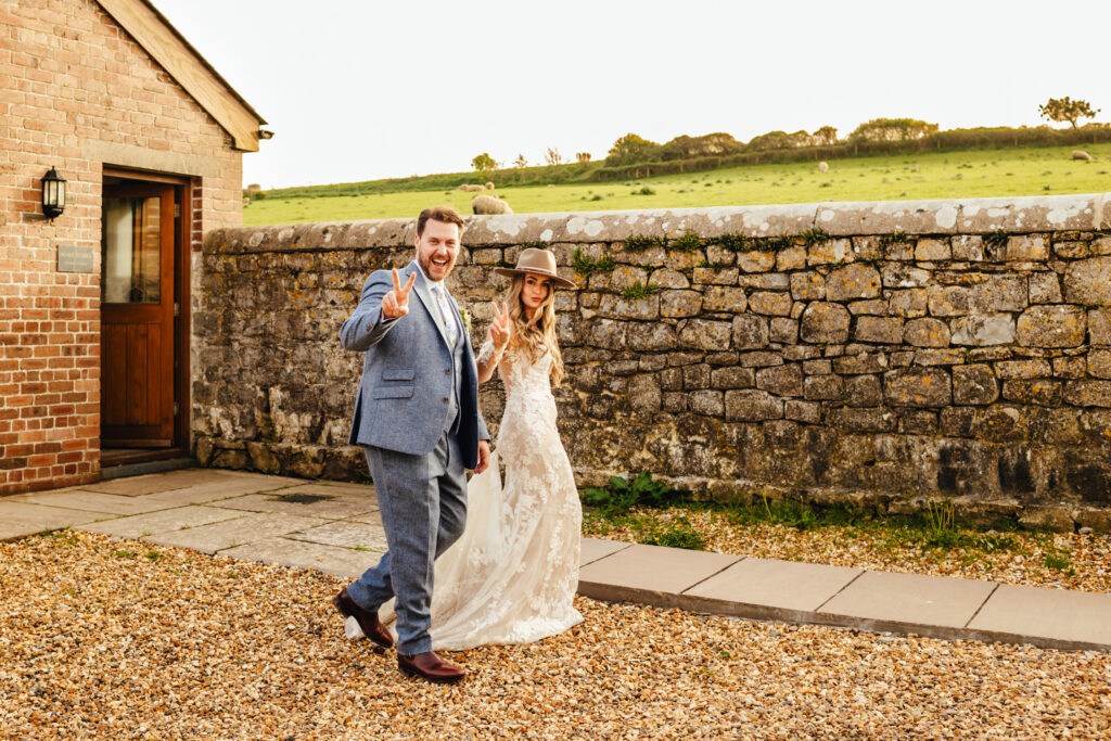 Bride in a lace dress and cowboy hat, and a groom in a blue suit walk past the photographer giving peace signs