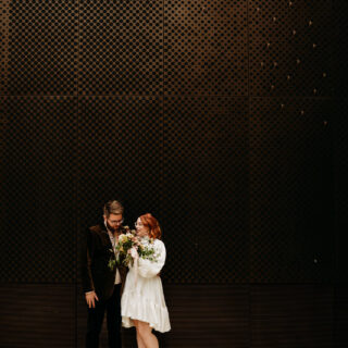Bride and groom in front of a plain wall smelling the bouquet