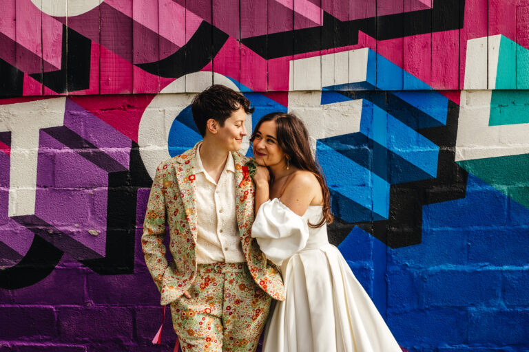 Two brides - one in a white dress and one in a floral suit - smile at each other in front of a colourful mural