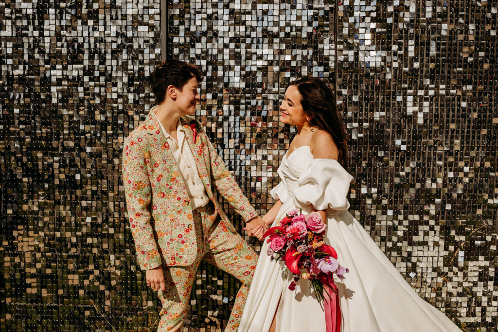 Two brides - one in a white dress and one in a floral suit - smile at each other in front of a silver shimmery backdrop