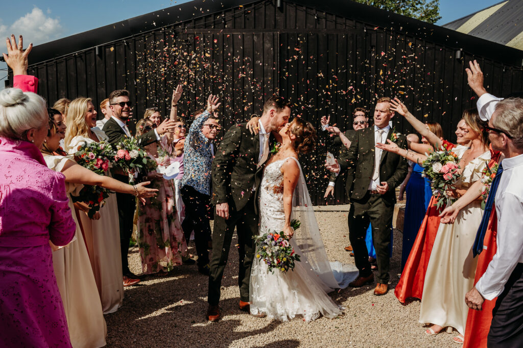A bride and groom kiss during the confetti walk, in front of a black barn