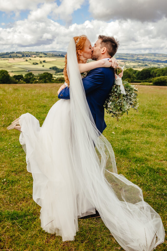 A fair skinned bride with long ginger hair is being picked up by a groom with brown hair and beard, they are kissing