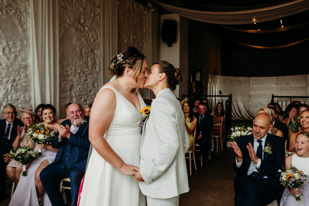 Two brides sharing their first kiss after getting married. One is wearing a floaty dress, the other is in a grey suit.