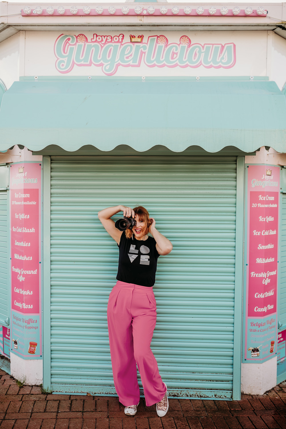 Cardiff Wedding Photographer - Charlie, a white woman with ginger hair, is holding a camera to her face and smiling, in front of a turquoise and pink building with a sign that reads 'Joys of Gingerlicious'