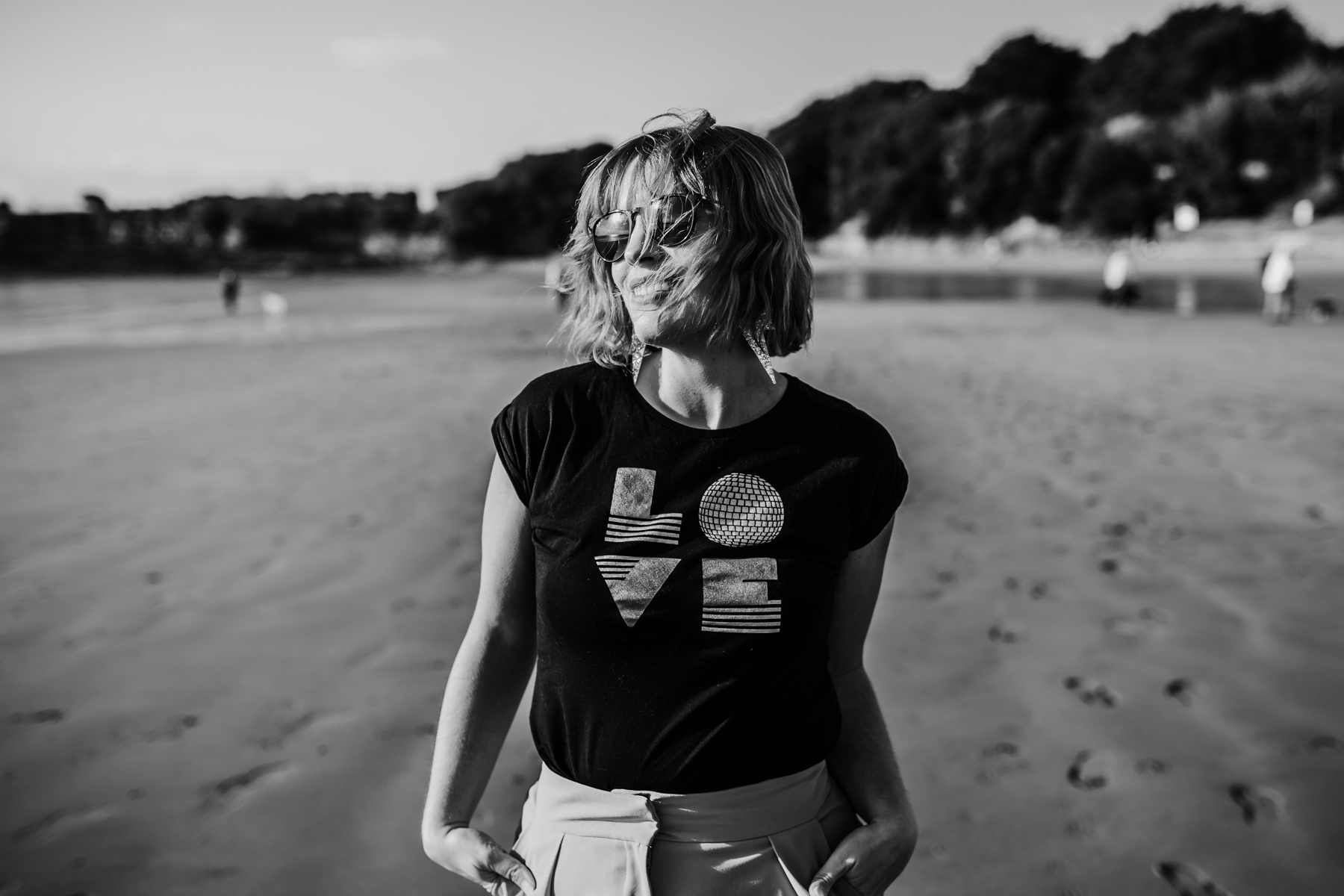 Black and white image of a white woman with a bob hairstyle and a T shirt that spells LOVE, on the beach, looking off towards the sun and smiling contentedly