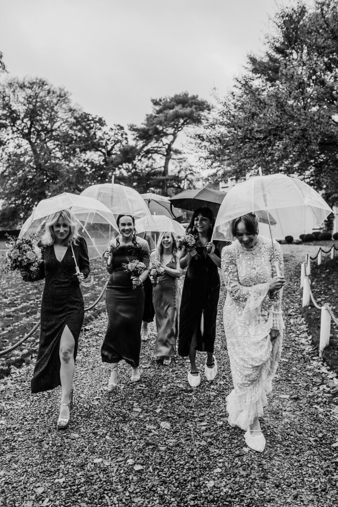 Black & White shot of a bride and her bridesmaids, walking along a path carrying umbrellas and wearing shoe covers in the rain