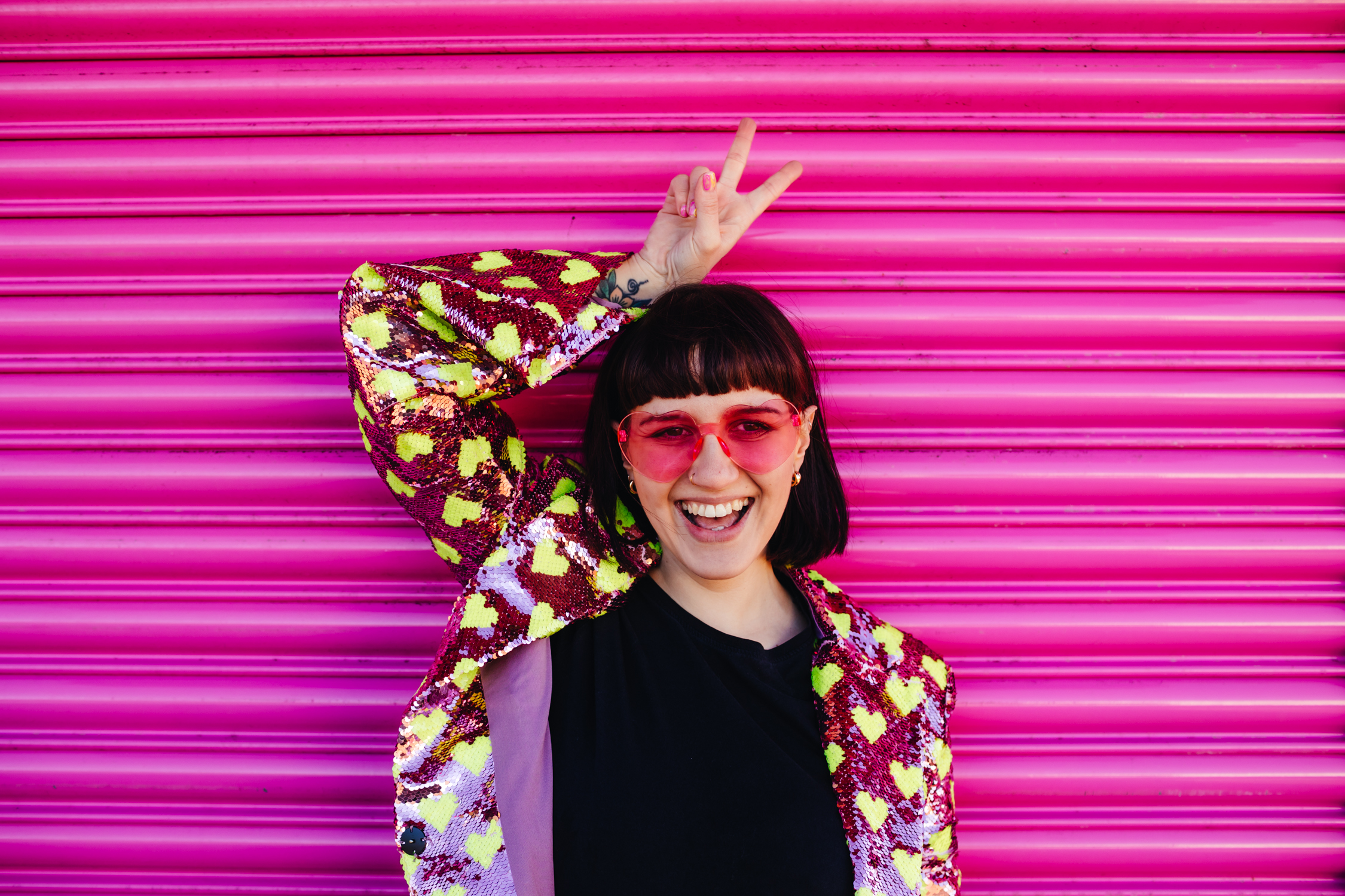 White woman with a funky haircut, pink heart shaped sunglasses and a sequin jacker standing in front of a bright pink shutter, holding her fingers in a peace sign and grinning