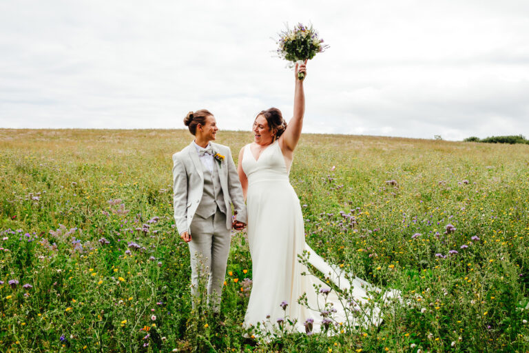 Two brides smiling happily at each other in a field. One has her bouquet high in the air