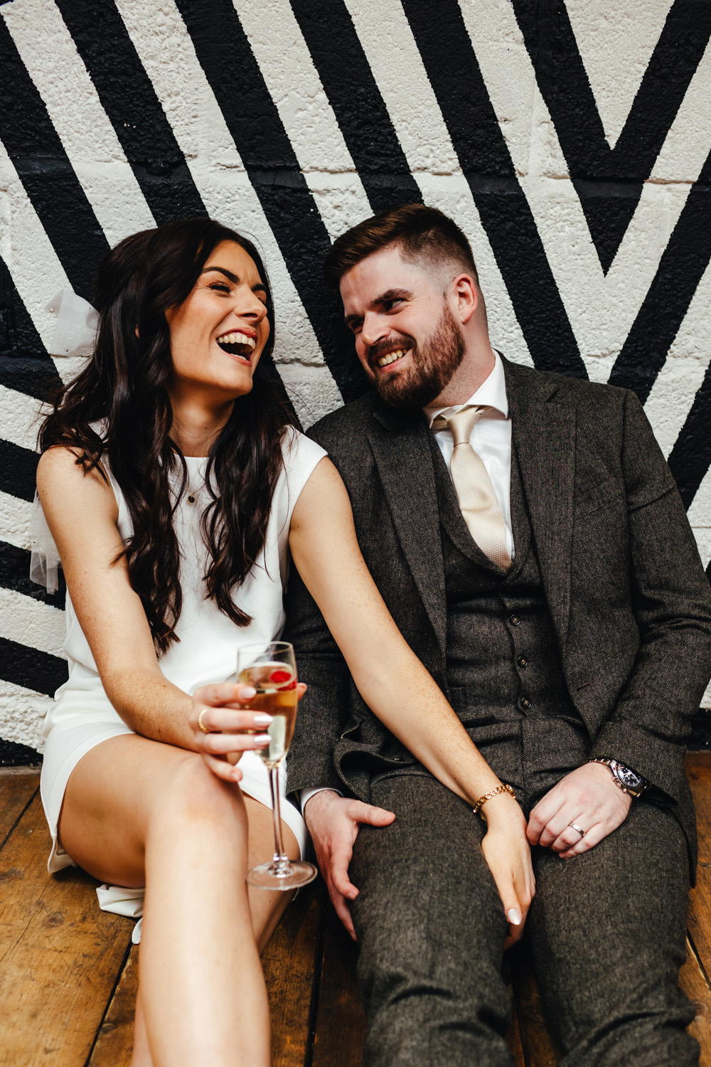 Bride & Groom sitting on the floor in front of a monochrome, geometric patterned wall. They are laughing and look relaxed