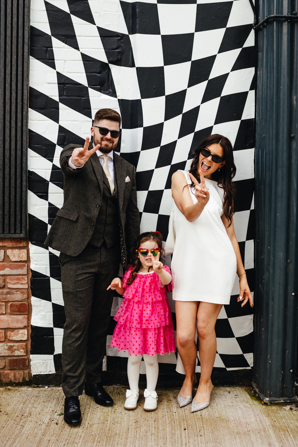 Wedding family portrait, bride in modern mini dress, grrom in a suit, little girl in a pink dress, standing in front of an abstract check wall, holding up their fingers in 'peace' signs