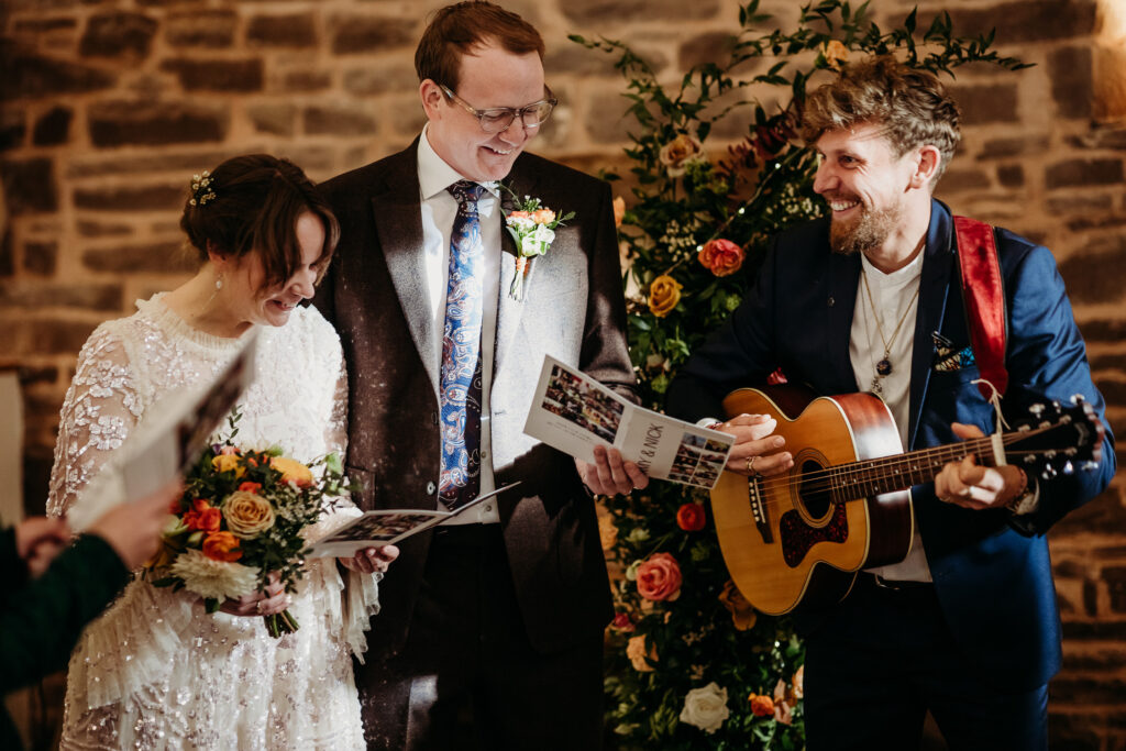 An acoustic guitar player is playing and smiling at the bride and groom singing next to him