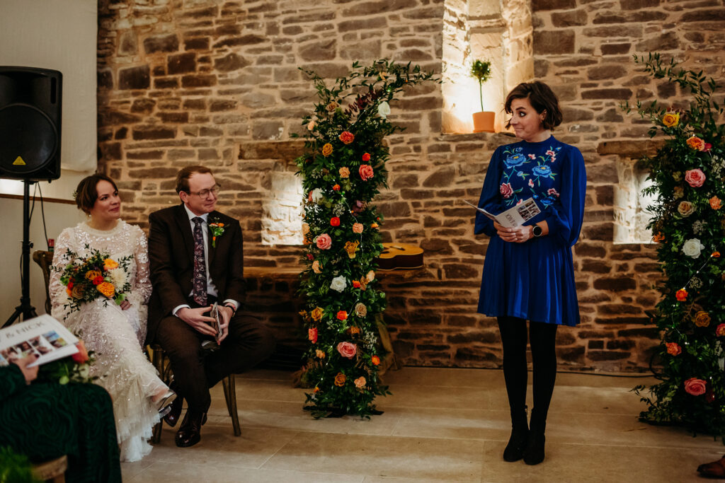 A bride and groom sit listening and watching their female friend perform a reading