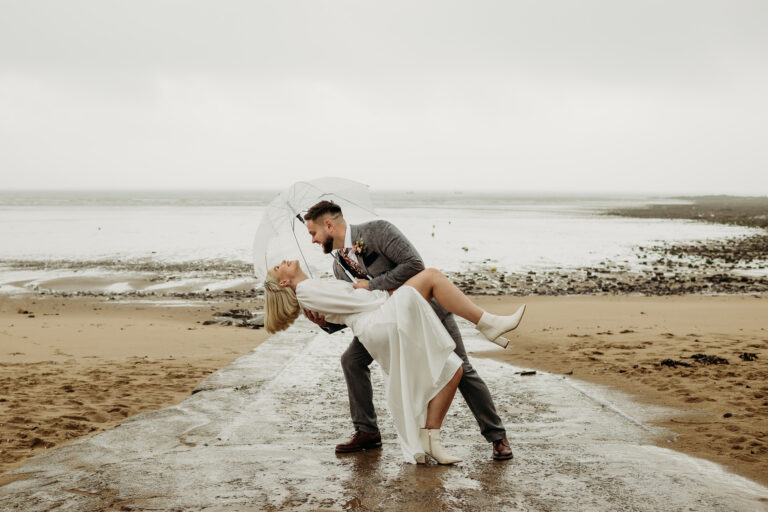 Bride and groom on a rainy beach. The groom is holding a clear umbrealla and 'dipping' the bride, who has a foot up in the air and is laughing