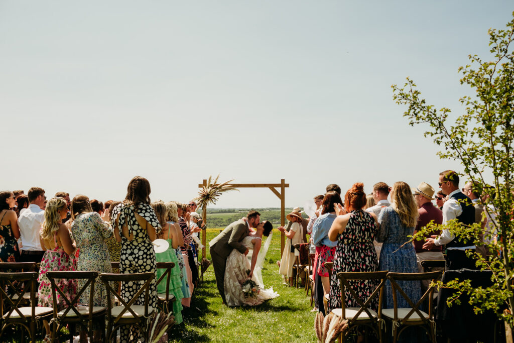 A bride and groom have a 'dip kiss' after their outdoor ceremony in front of their guests and a wooden archway