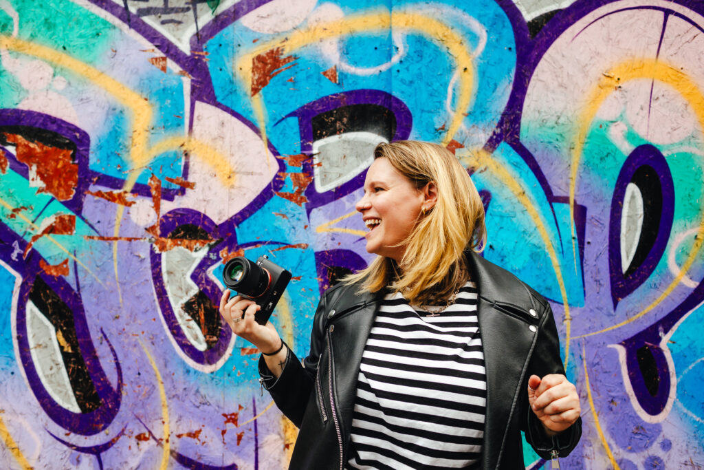 A white woman with blonde hair is holding a camera casually, looking off to her right and laughing, while stood in front of purple and blue graffiti