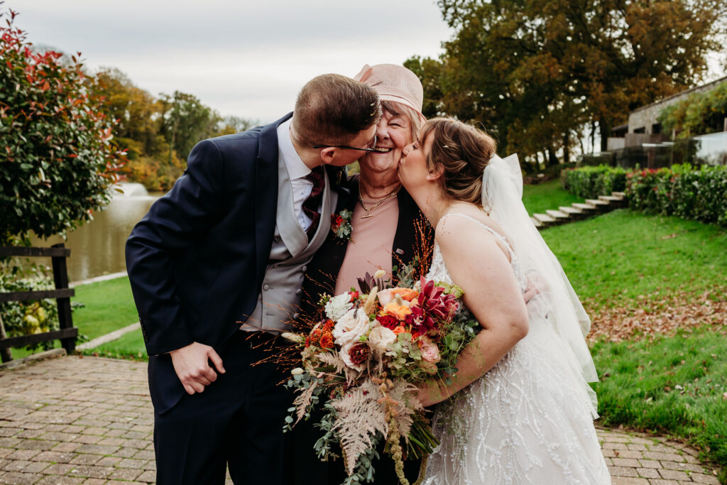 An older lady stands in the middle of a bride and groom while they both kiss her on the cheeks