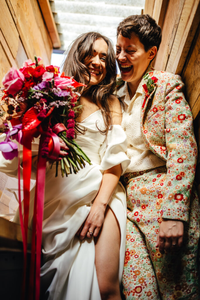 Two brides - one in a white dress and one in a floral suit - are squeezed together in the narrow staircase of a double decker bus, laughing