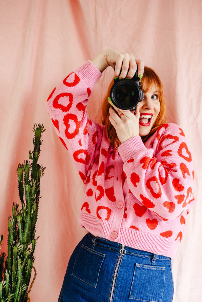 White female with ginger hair, wearing a bright pink cardigan with red hearts on and a denim skirt, stands in front of a mid pink backdrop next to a plant, holding her camera up to her face and smiling