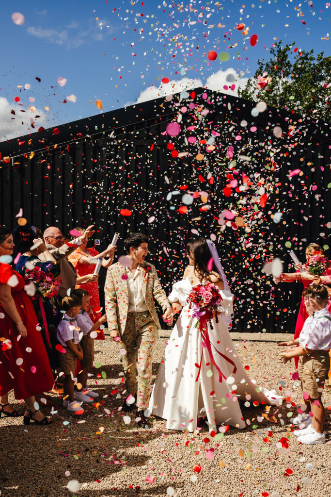 Two brides - one on a dress and one in a floral suit - in an epic confetti walk with tonnes of pink and red paper confetti in the air