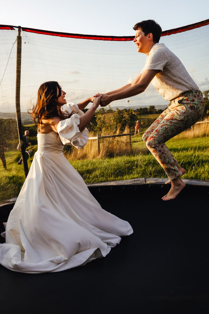 Two brides - one in a white dress and one in a floral suit - jumping on a trampoline