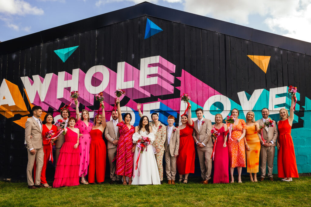 A wedding group photo in front of a big black barn with a colourful mural that says "a whole lot of love". The couple consists of a bride with long hair in a white dress, a bride with short hair in a floral suit, and a wedding party wearing a mix of pink, red and orange (women) and beige suits (men)