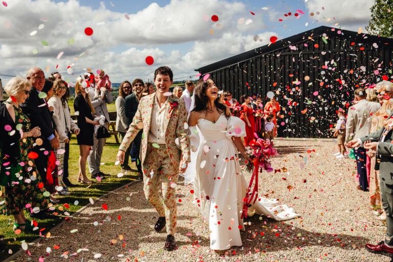 A bride in a floral suit and a bride in a big white dress walk hand in hand through a shower of red and pink paper confetti, lined by wedding guests in front of a black barn