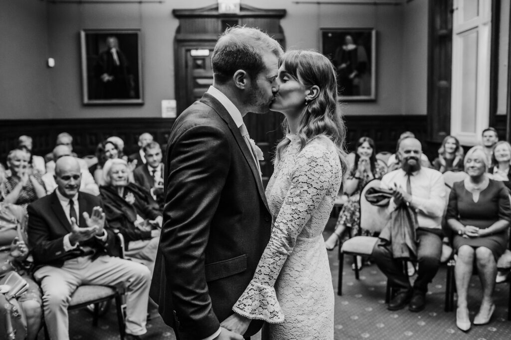 Black and white photograph of a newlywed couple sharing their first kiss in a register office in front of their guests