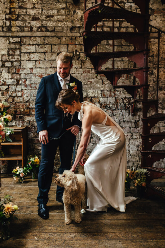 A bride in a satin dress bending down to pat her  golden cockapoo, mid ceremony, while the groom watches on, smiling