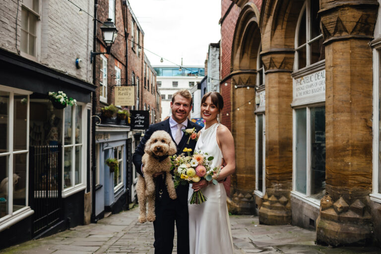 Bride and Groom couple photo with their golden cockapoo at the Christmas Steps. The bride is wearing a white satin dress, the groom in a black suit.