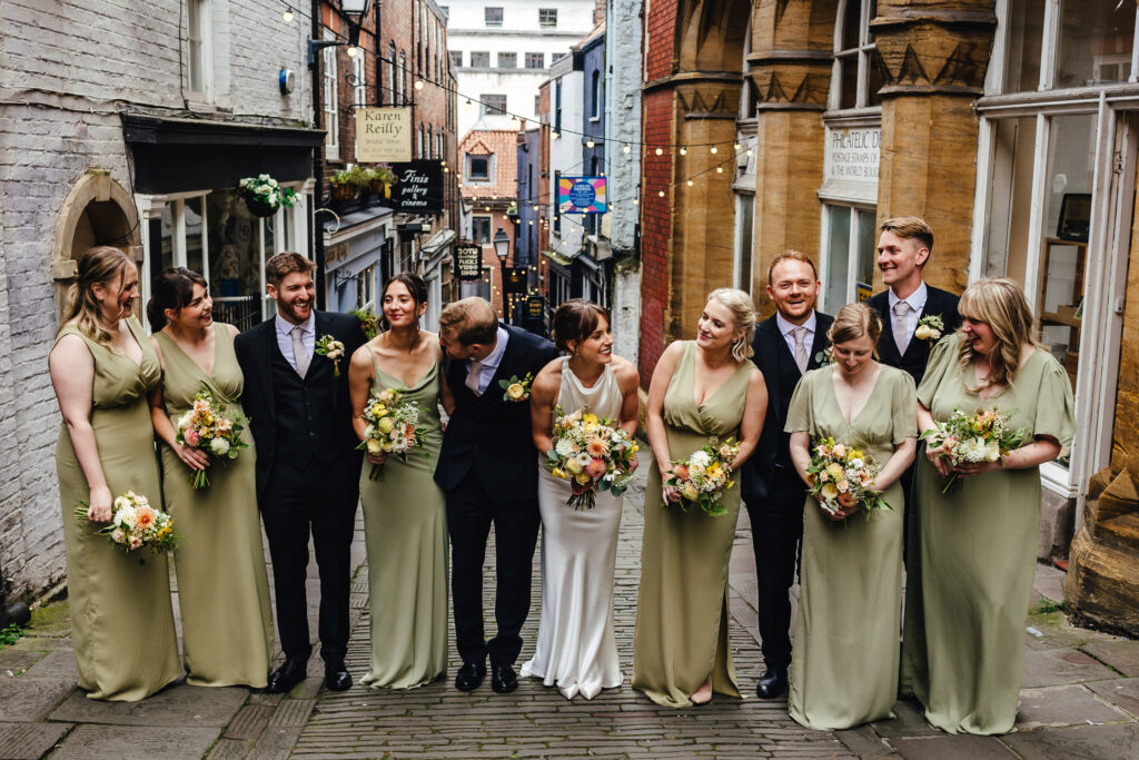 Group wedding photo at the Christmas steps. The bride wears a white satin dress, then men are all in dark suits and the qomen in long sage green dresses