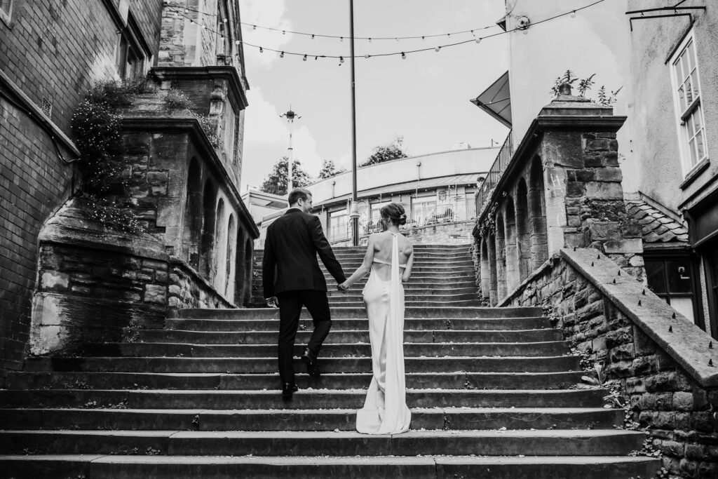 Black and white image of a glamorous bride and groom walking away from the camera, up some steps, holding hands