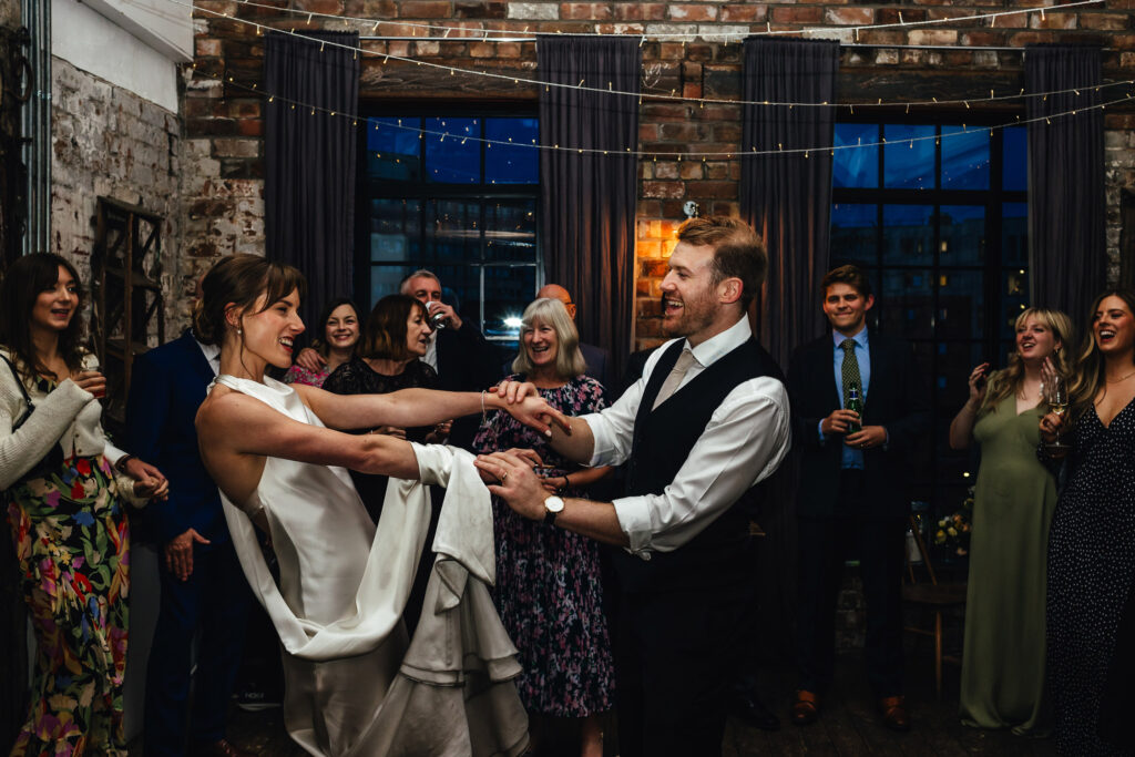 Bride and Groom in their 'first dance' in front of their wedding guests in a red brick, urban venue with lots of string lights