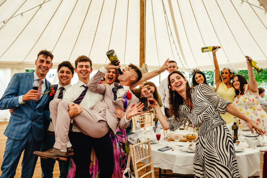 A fun group photo at a table in a marquee, one of the grooms is being carried while he drinks from a wine bottle