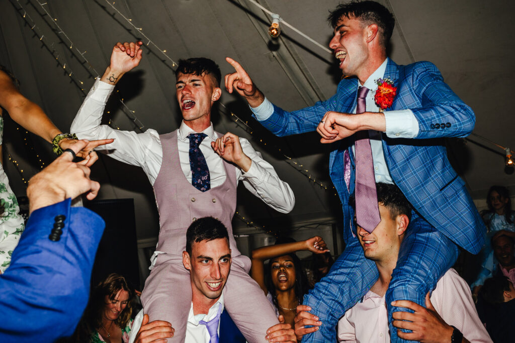 A dancefloor at a wedding, two grooms are on the shoulders of two of their friends, hands in the air