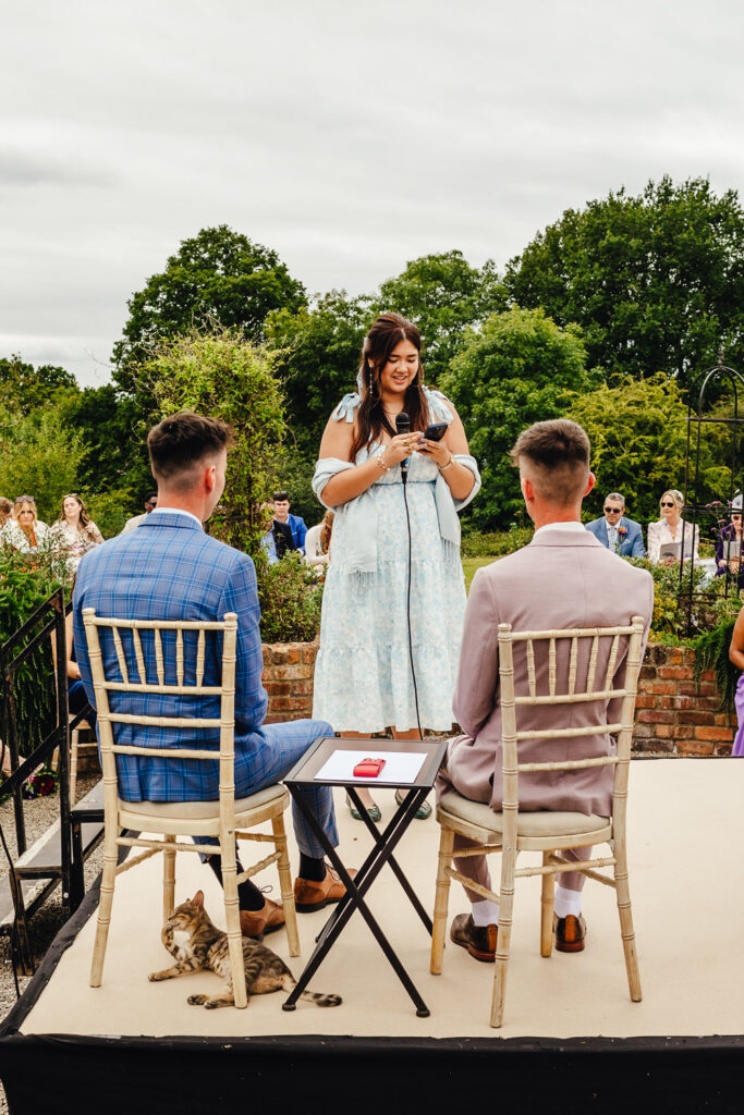 A light brown-skinned woman is reading to two grooms during an outdoor wedding ceremony, while a cat cleans it's paw underneath one of their chairs