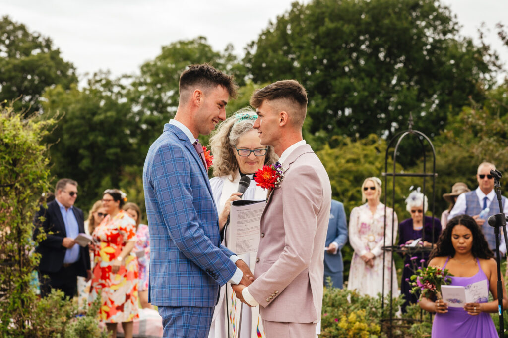 Two grooms - one in pink and one in blue - being married outdoors by a female vicar