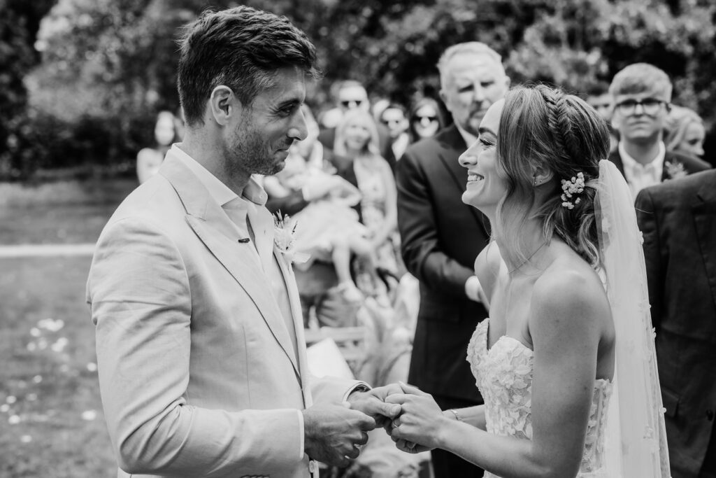 Black & White image of a bride and groom smiling at each other during their wedding ceremony, the groom is holding the bride's left hand.