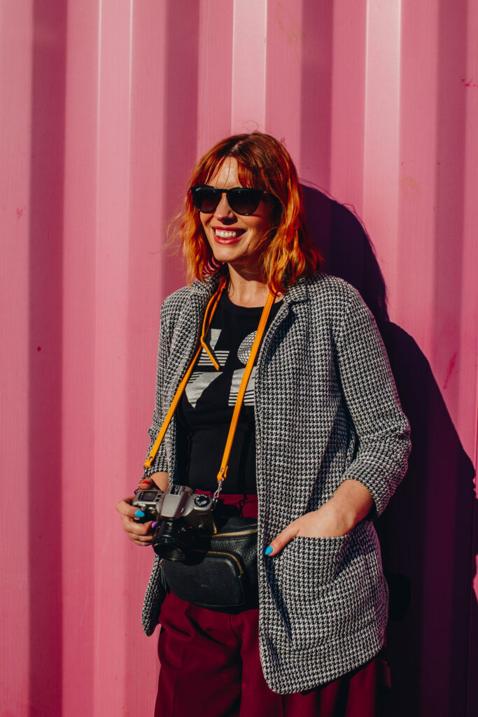 A woman with ginger hair leaning against a pink corrugated background holding a camera on a yellow strap