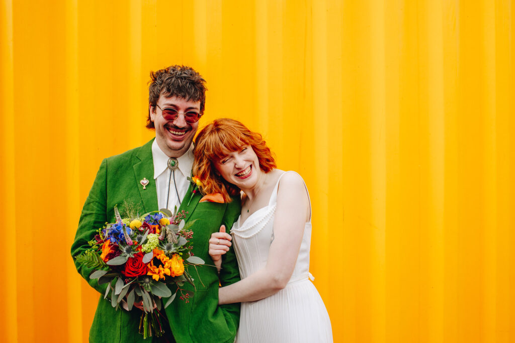 A groom holding a bouquet and a bride leaning her head on his shoulder, in front of a yellow corrugated wall, looking off camera and laughing.