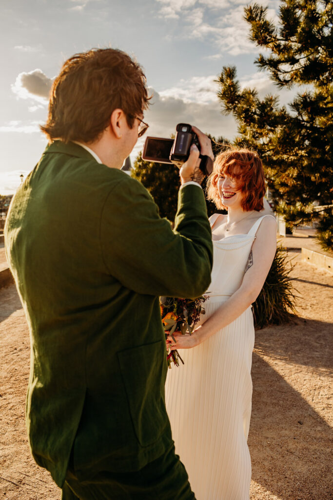 A bride with ginger hair laughing, while a groom films her with an old school camcorder